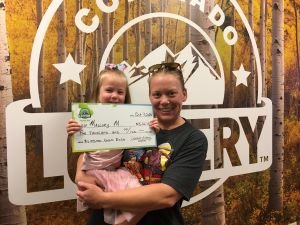 Mallory M. y su hija sosteniendo un cheque de gran tamaño por $5,000 frente al fondo de la Lotería de Colorado