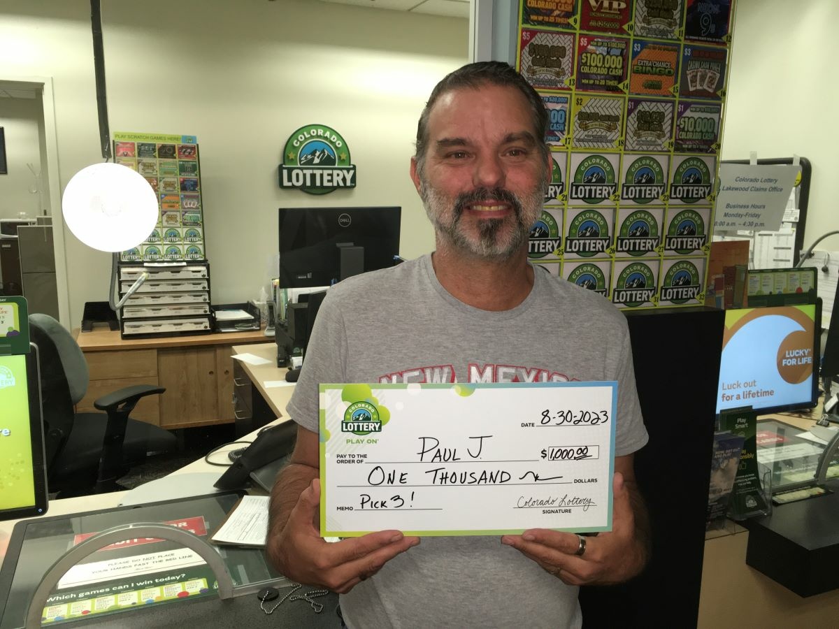 Paul J. of Denver holding an oversized check for $1,000 in front of Colorado Lottery background.