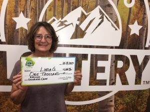 Linda G sosteniendo un cheque de gran tamaño por $1,000 frente al fondo de la Lotería de Colorado
