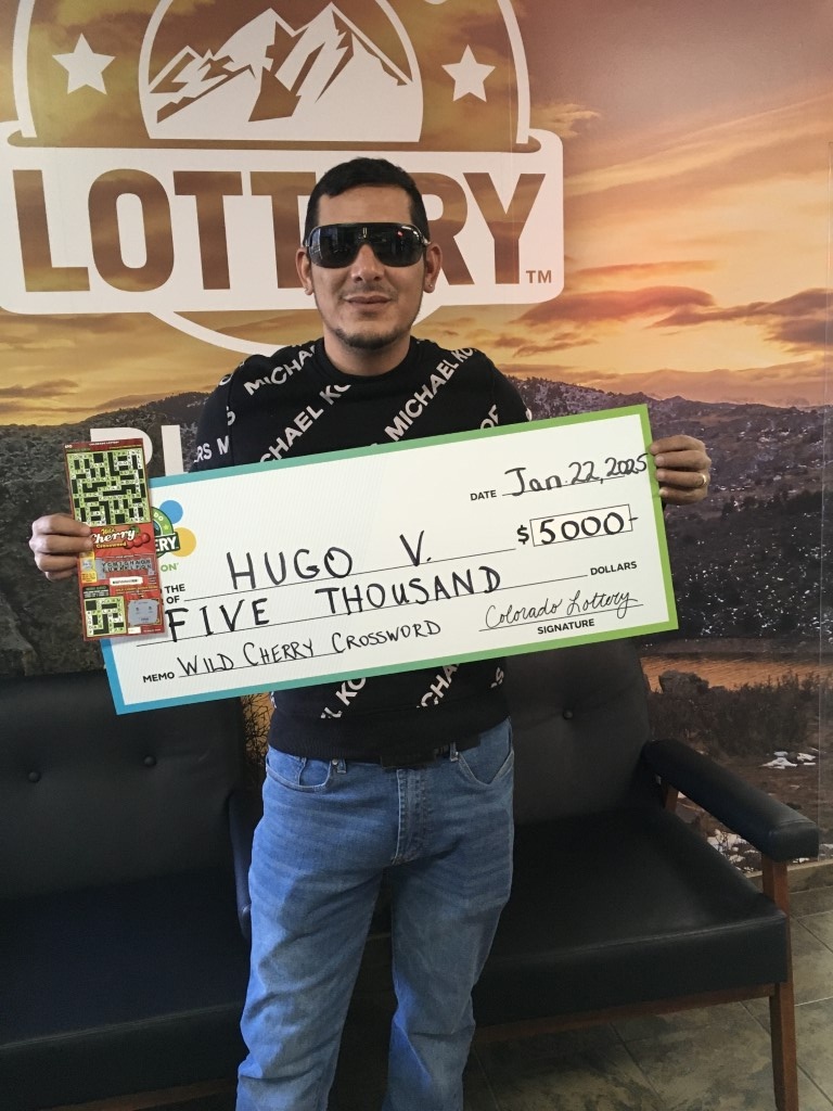 Hugo V. sosteniendo un cheque por $100,000 frente al fondo de la Lotería de Colorado.