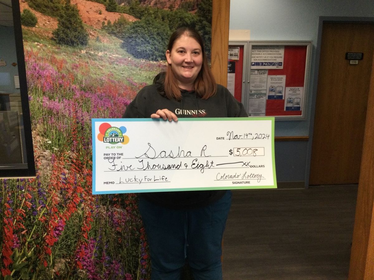 Sasha R. sosteniendo el cheque gigante de $5,000 frente al fondo de Colorado Lottery