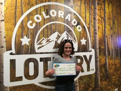 Deena T. holding an oversized check for $1,000 in front of Colorado Lottery background