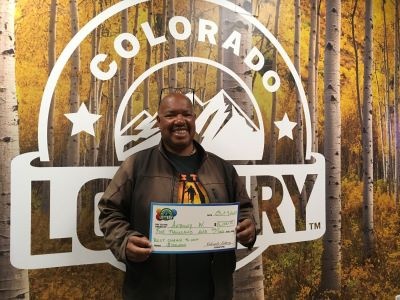 Anthony W. sosteniendo un cheque gigante por $5,000 frente al fondo de la Lotería de Colorado