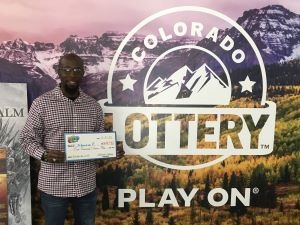 Khourie F. sosteniendo un cheque gigante por $5,000 frente al fondo de la Lotería de Colorado