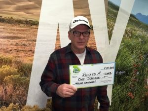 Richard M. sosteniendo el cheque de $1,000 frente al fondo de Colorado Lottery