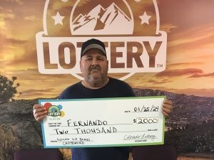 Fernando T. sosteniendo un cheque de gran tamaño por $2,000 frente al fondo de la Lotería de Colorado