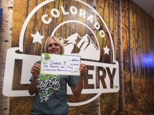 Sandra P. sosteniendo un cheque gigante de $5,000 frente al fondo de la Lotería de Colorado.