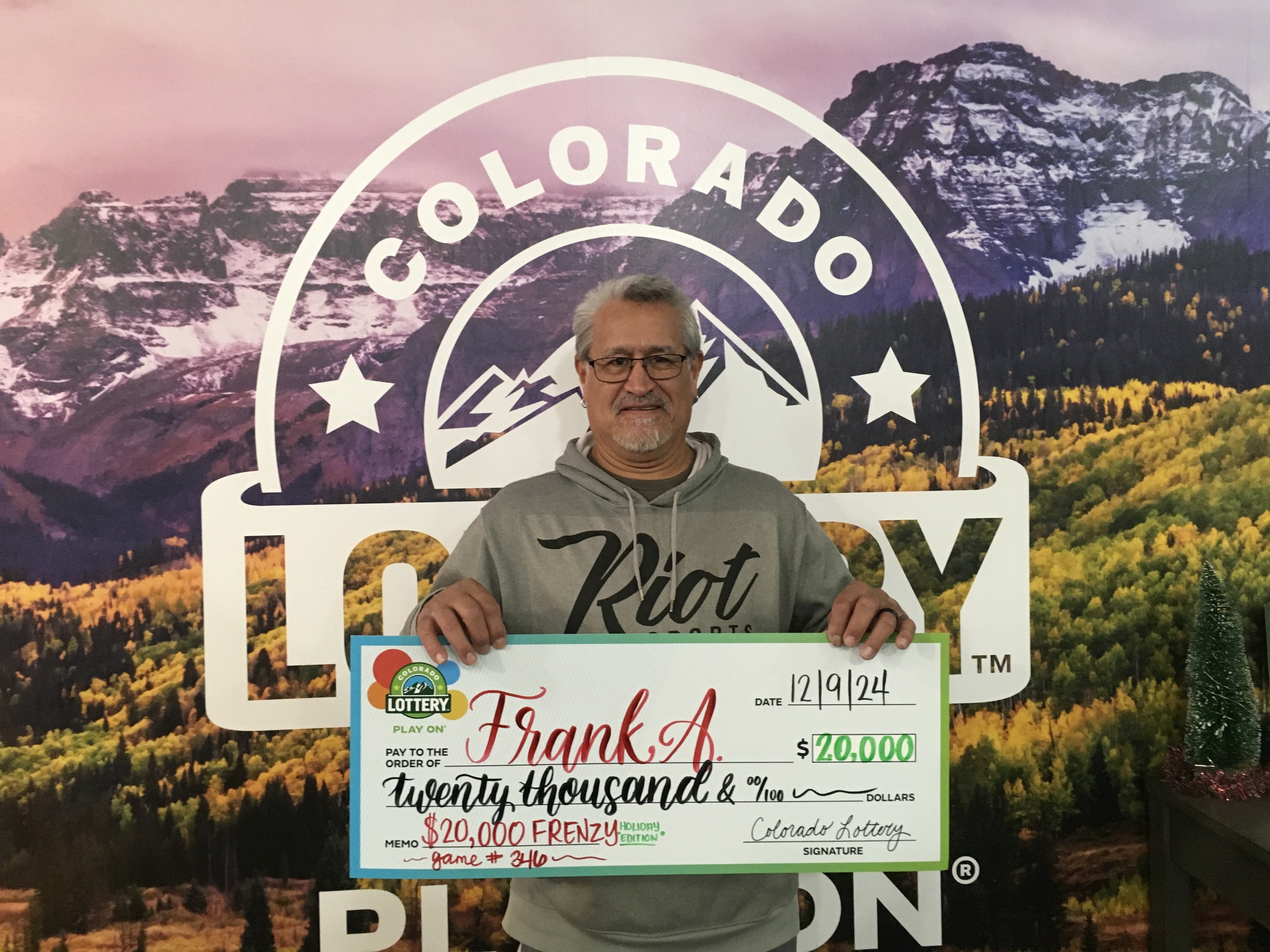 Frank A. sosteniendo el cheque gigante de $20,000 frente al fondo de Colorado Lottery