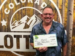 Mike G. de Longmont sosteniendo un cheque de gran tamaño por $1,000 frente al fondo de la Lotería de Colorado.