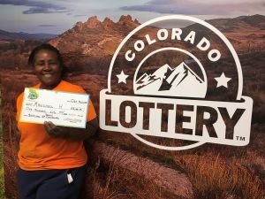 Mauchell H. sosteniendo un cheque de gran tamaño por $1,000 frente al fondo de la Lotería de Colorado