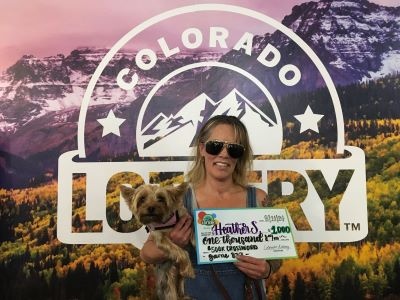 Heather S. holding an oversized check for $1,000 in front of Colorado Lottery background