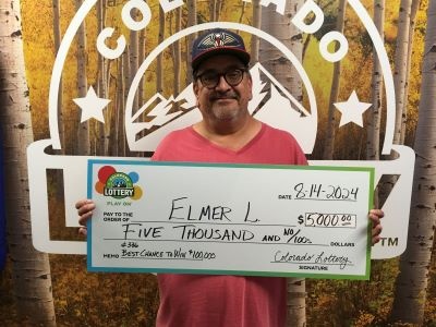 Elmer L. sosteniendo un cheque gigante de $5,000 frente al fondo de la Lotería de Colorado