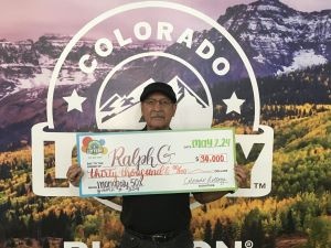 Ralph G. sosteniendo un cheque gigante por $30,000 frente al fondo de la Lotería de Colorado