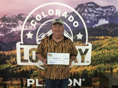 Herbert E. sosteniendo un cheque gigante de $1,000 frente al fondo de la Lotería de Colorado