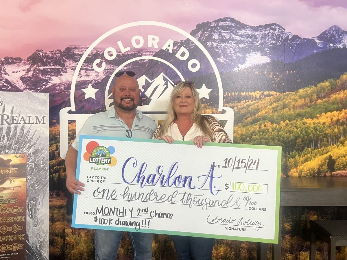 Charlon A. sosteniendo un cheque gigante de $100,000 frente a un fondo de la Lotería de Colorado.