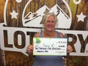 Carole V. de Littleton sosteniendo un cheque de gran tamaño por $2,500 frente al fondo de la Lotería de Colorado