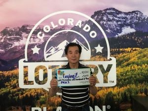 Geying Z. sosteniendo un cheque de gran tamaño por $5,600 frente al fondo de la Lotería de Colorado