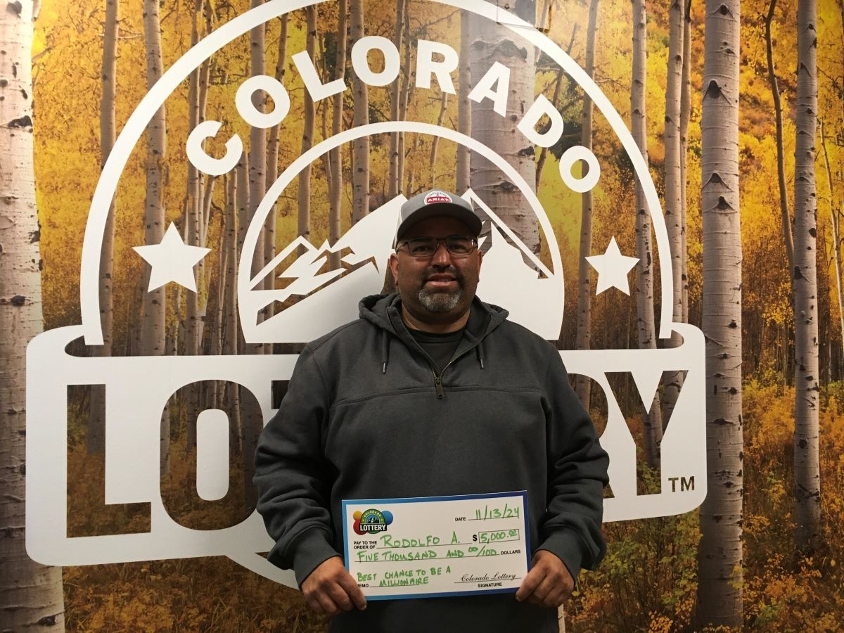 Rodolfo A. sosteniendo el cheque gigante de $5,000 frente al fondo de Colorado Lottery