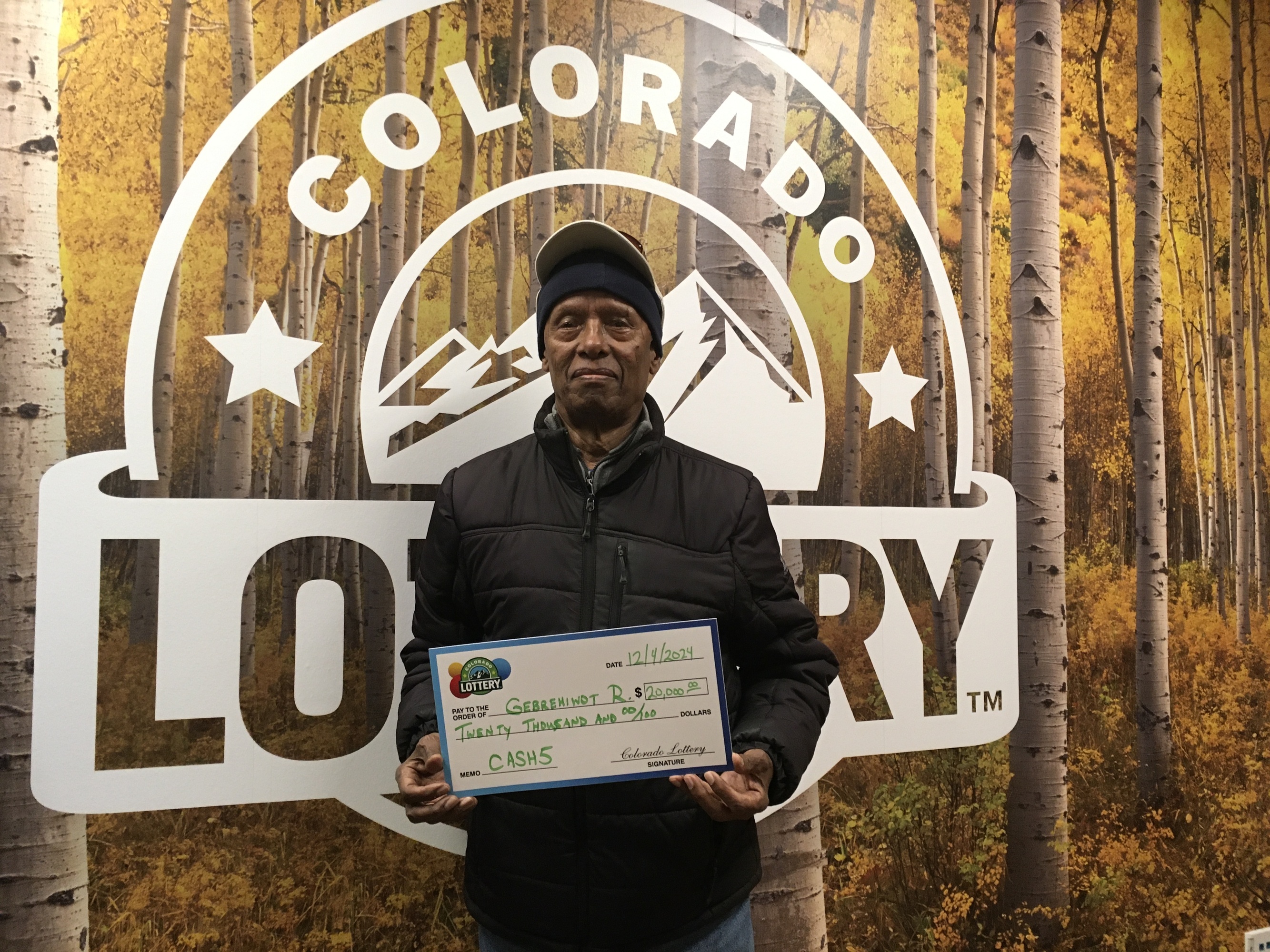 Gebrehiwot R. sosteniendo el cheque gigante de $20,000 frente al fondo de Colorado Lottery