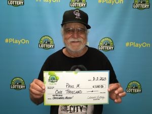Paul M. de Littleton sosteniendo un cheque de gran tamaño por $1,000 frente al fondo de la Lotería de Colorado.