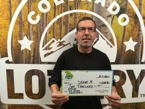 Shane M. de Arvada sosteniendo un cheque de gran tamaño por $2,000 frente al fondo de la Lotería de Colorado