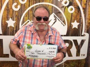 Victor W. de Arvada sosteniendo un cheque de gran tamaño por $2,000 frente al fondo de la Lotería de Colorado.