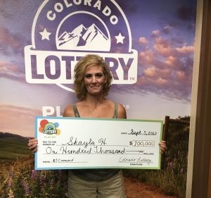 Shayla H. de Mack sosteniendo un cheque de gran tamaño por $100,000 frente al fondo de la Lotería de Colorado.