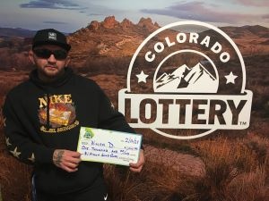 William D. sosteniendo un cheque de gran tamaño por $1,000 frente al fondo de la Lotería de Colorado