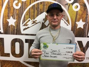 Scott S. sosteniendo un cheque de gran tamaño por $1,000 frente al fondo de la Lotería de Colorado